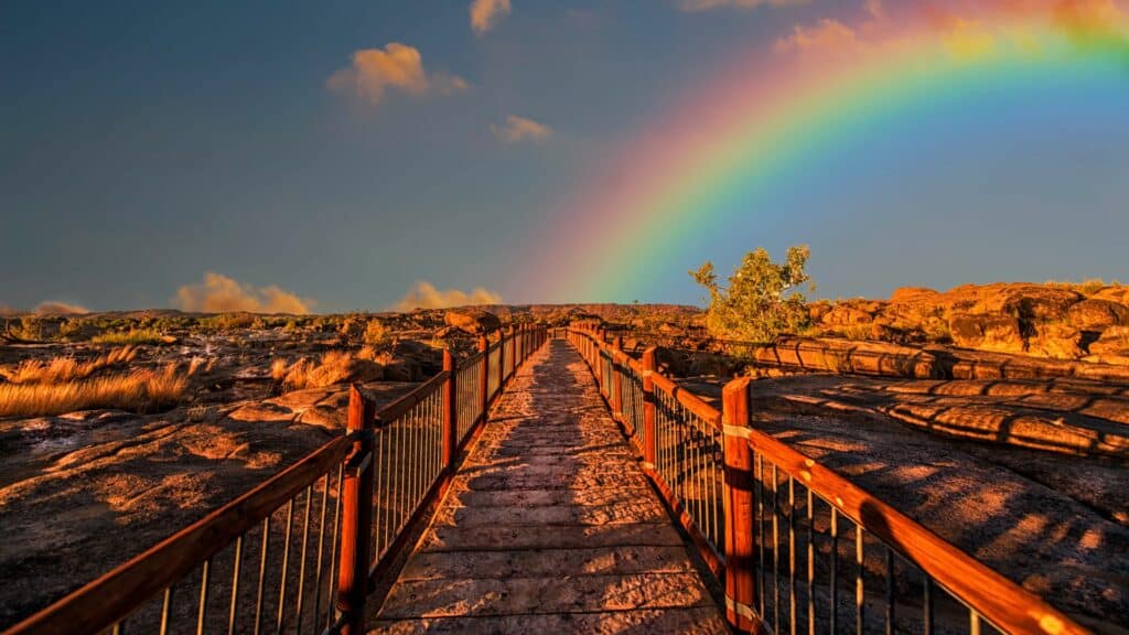 Imagem de uma ponte de madeira no meio de umas pedras no chão, em uma paisagem bonita e um arco-iris no céu