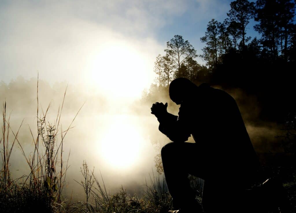 Silhueta de homem ajoelhado, com as mãos juntas em oração, em meio à natureza. Há neblina à sua frente.