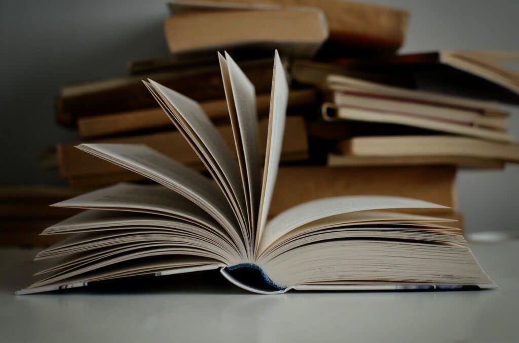 Livro aberto, com as folhas em movimento, sobre uma superfície branca. Atrás dele há uma pilha de outros livros.