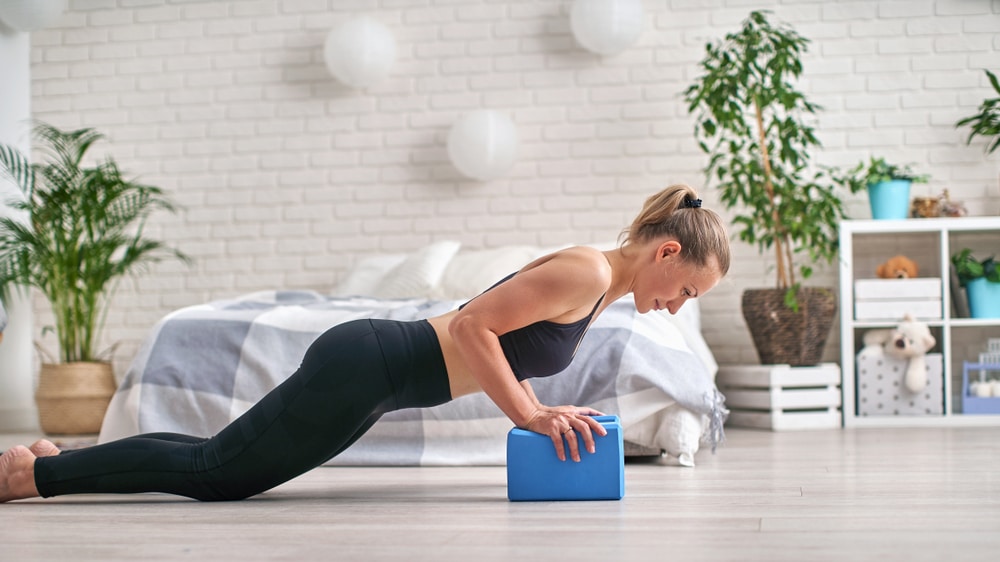 Mulher fazendo Yoga com um bloco azul dentro de seu quarto