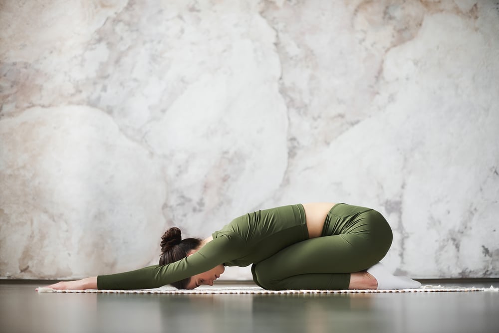 Mulher realizando a postura balasana da Yoga em ambiente fechado