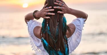 Mulher de frente para o mar usando uma bata branca e acessários azuis como penas e anéis