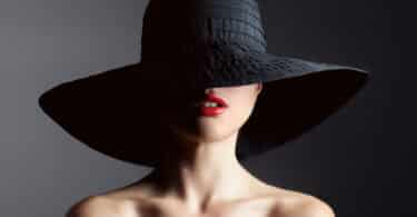Mulher glamurosa de ombros nus, batom vermelho e um grande chapéu preto cobrindo metade de seu rosto