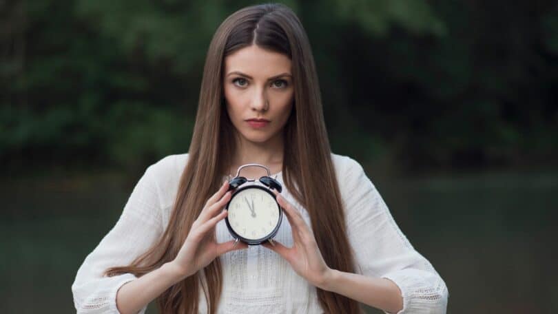 Imagem de uma mulher segurando um relógio na sua frente e olhando com semblante sério para a tela