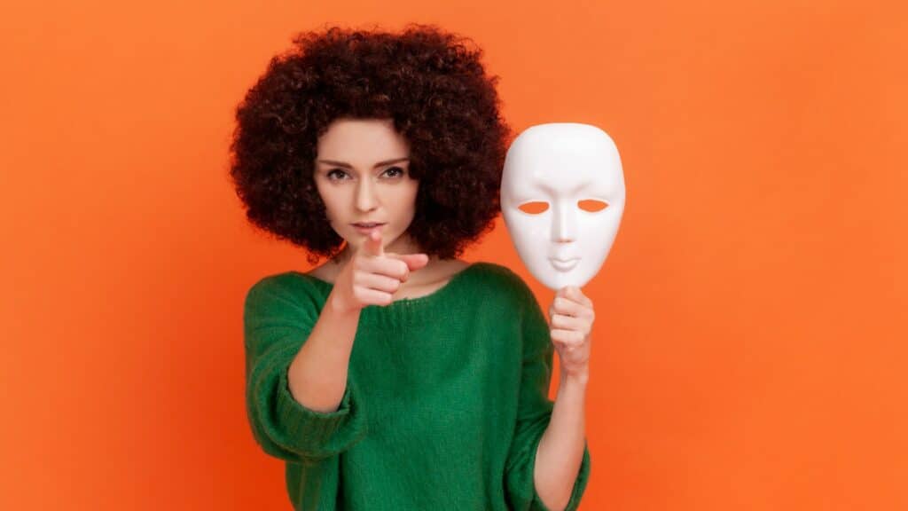 Imagem de uma mulher em um fundo laranja olhando para a tela e apontando com uma mão e com a outra segura uma máscara