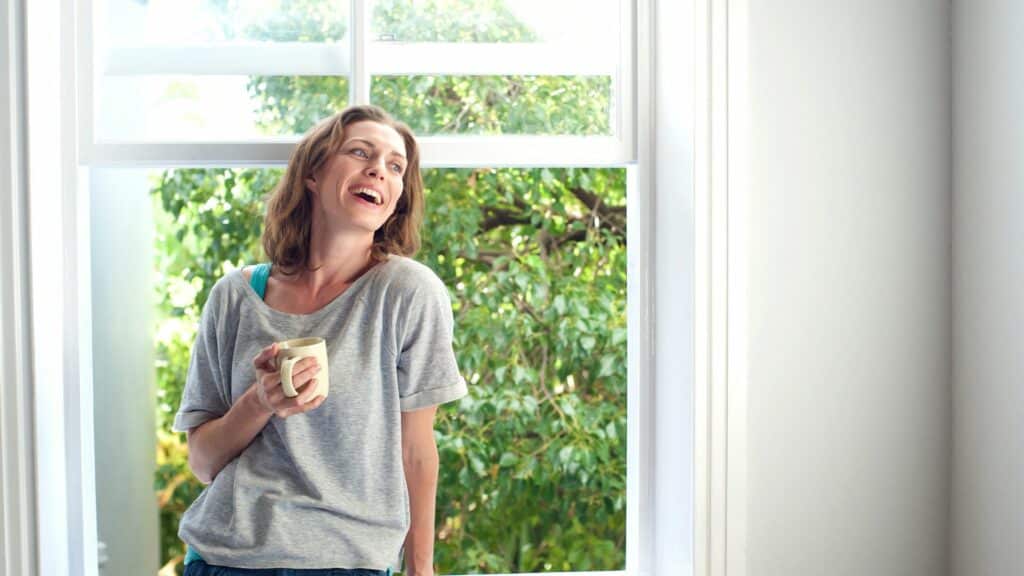 Imagem de uma mulher sorrindo encostada em uma janela e ao fundo é possível ver as árvores lá fora