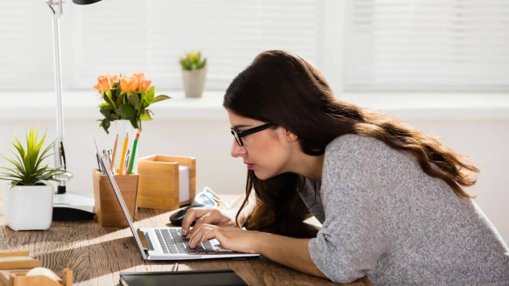 Imagem de uma mulher debruçada sobre a mesa em frente ao laptop, com uma postura nada favorável