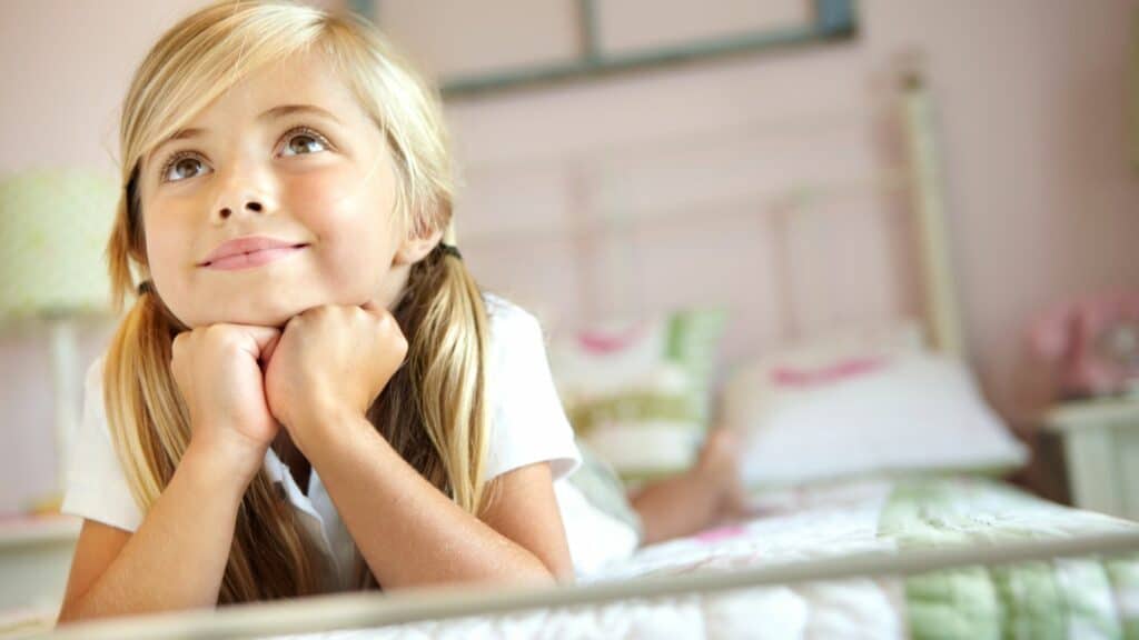 Imagem de uma criança em sua cama com as mãos no queixo e sorrindo, como se estivesse sonhando acordada