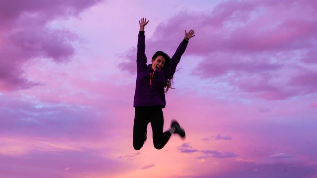 Imagem de uma mulher pulando e com os braços erguidos e sorrindo e atrás dela o céu arroxeado