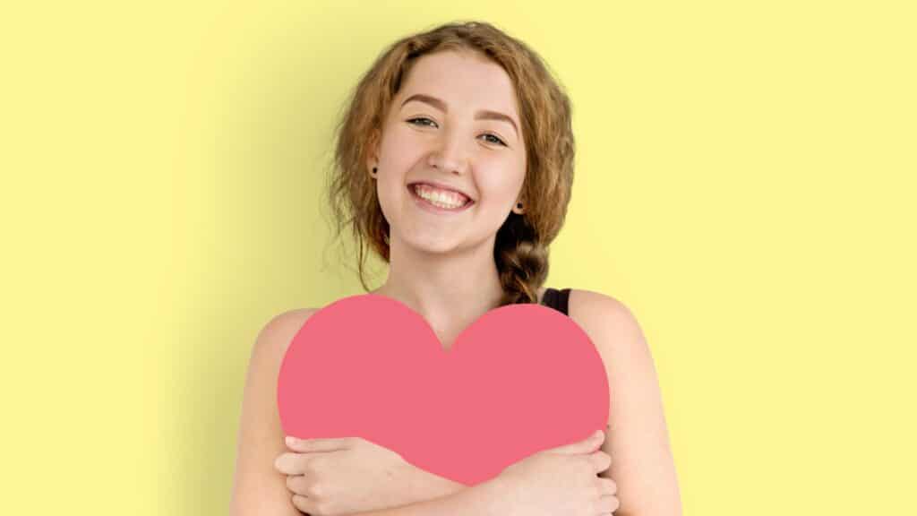 Imagem de uma moça segurando um coração, que parece de papel, está sorrindo em um funso amarelo