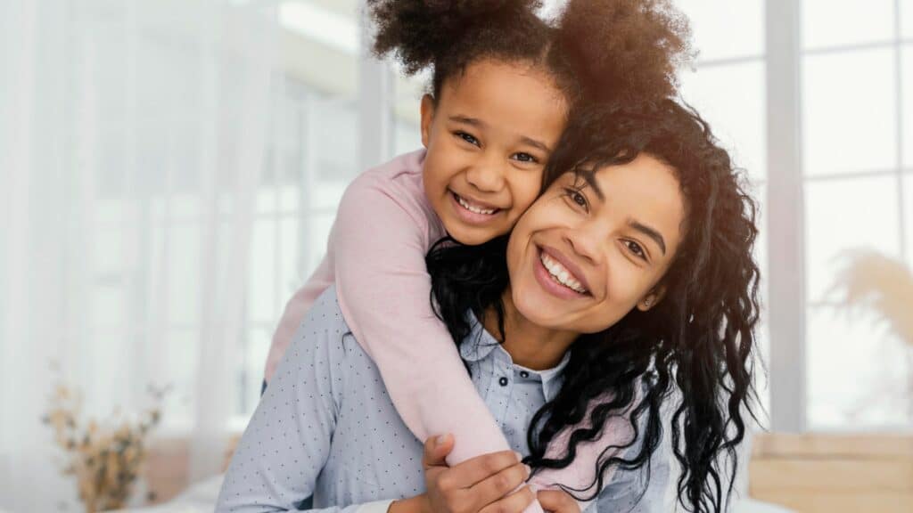 Imagem de uma mãe com sua filha abraçando ela no pescoço e ambas estão sorrindo