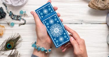 Mãos femininas segurando cartas de Tarot em cima de uma mesa de madeira com cristais e outros utensílios místicos