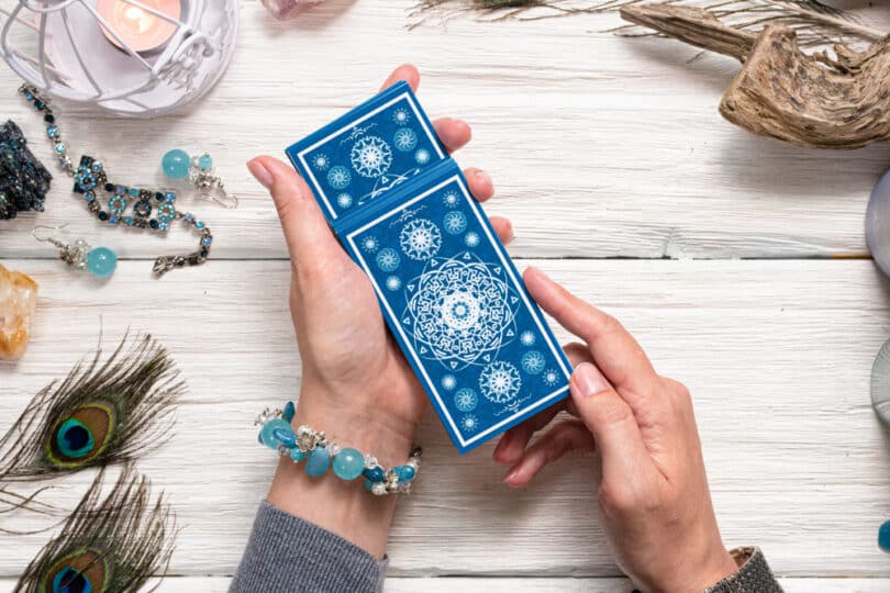 Mãos femininas segurando cartas de Tarot em cima de uma mesa de madeira com cristais e outros utensílios místicos