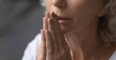 Mulher idosa com as mãos juntas perto do rosto e os olhos fechados, rezando