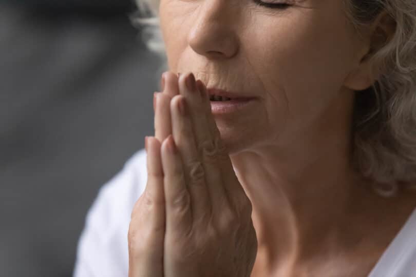 Mulher idosa com as mãos juntas perto do rosto e os olhos fechados, rezando