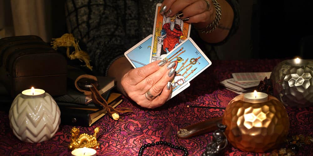Mãos de uma mulher segurando cartas de Tarot em uma mesa cheia de itens místicos