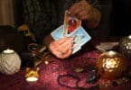 Mãos de uma mulher segurando cartas de Tarot em uma mesa cheia de itens místicos