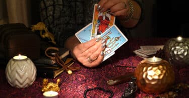 Mãos de uma mulher segurando cartas de Tarot em uma mesa cheia de itens místicos