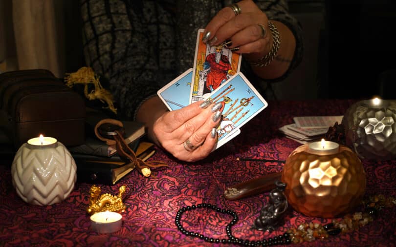 Mãos de uma mulher segurando cartas de Tarot em uma mesa cheia de itens místicos