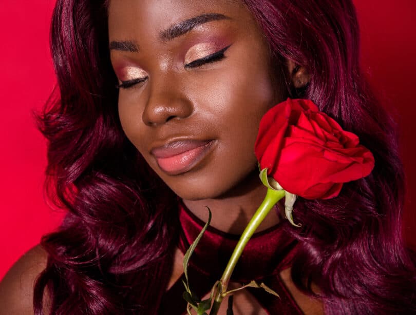 Mulher maquiada em tons de vermelho segurando uma rosa vermelha de olhos fechados