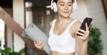 Mulher em roupas esportivas utilizando fones de ouvido, segurando celular e tapete de Yoga