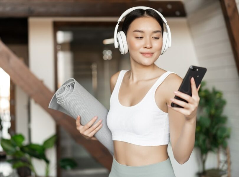 Mulher em roupas esportivas utilizando fones de ouvido, segurando celular e tapete de Yoga