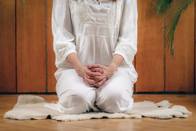 Mulher sentada sobre os calcanhares fazendo Kriya Yoga em ambiente interno