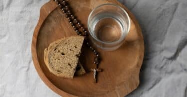 Um copo de água e um pedaço de pão sobre tábua de madeira com um terço religioso