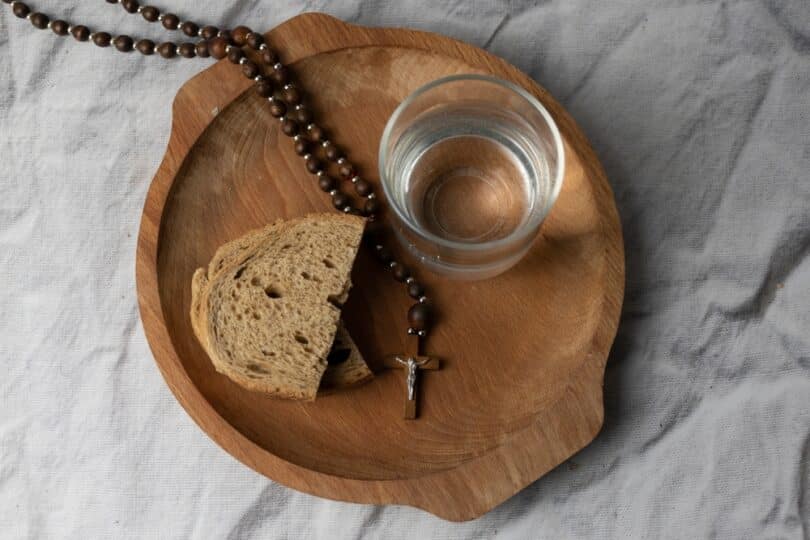 Um copo de água e um pedaço de pão sobre tábua de madeira com um terço religioso