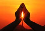 Silhueta de duas mãos juntas com um raio de sol passando pelo centro delas, em sinal de oração