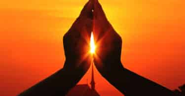 Silhueta de duas mãos juntas com um raio de sol passando pelo centro delas, em sinal de oração