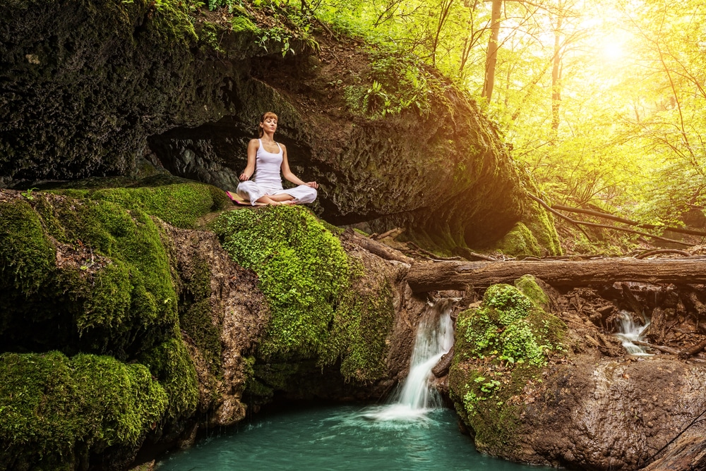 Mulher meditando em cima de uma pedra no meio da natureza. Ao lado dela, há uma mini-cachoeira