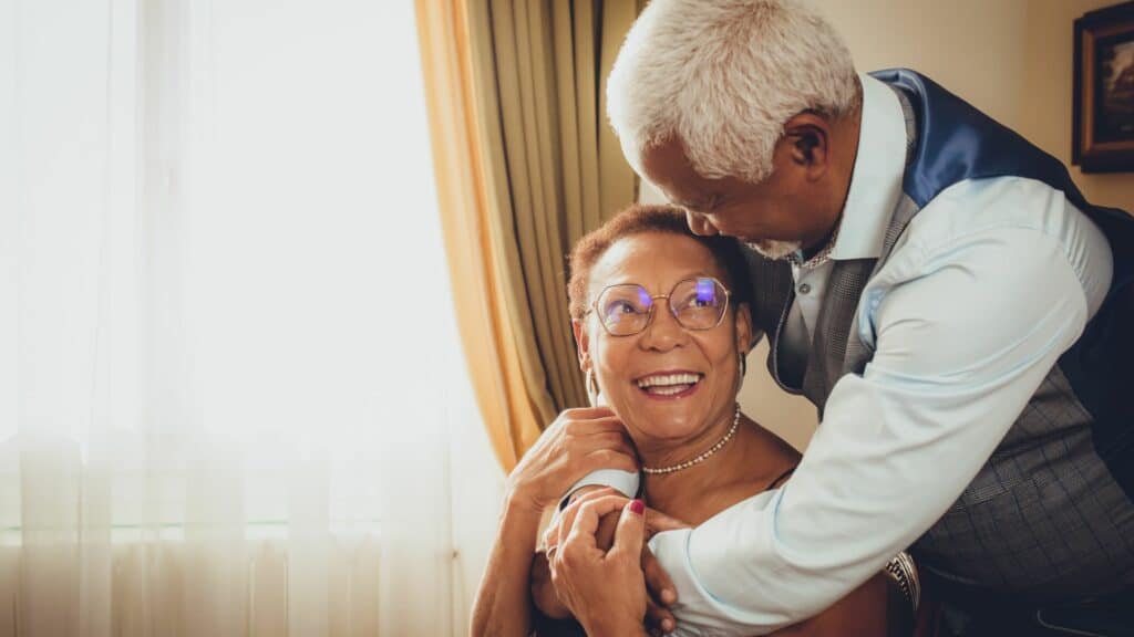 Senhor idoso abraçando sua esposa enquanto os dois se olham e sorriem. 