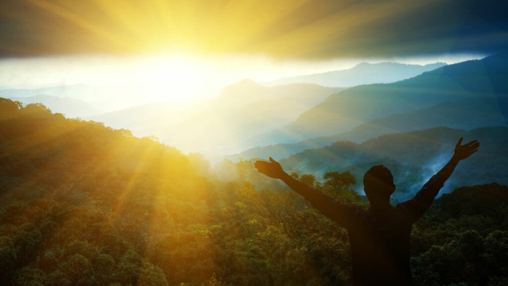 Imagem de um lindo pôr-do-sol em uma montanha em que um homem está com os braços abertos e estendidos para cima