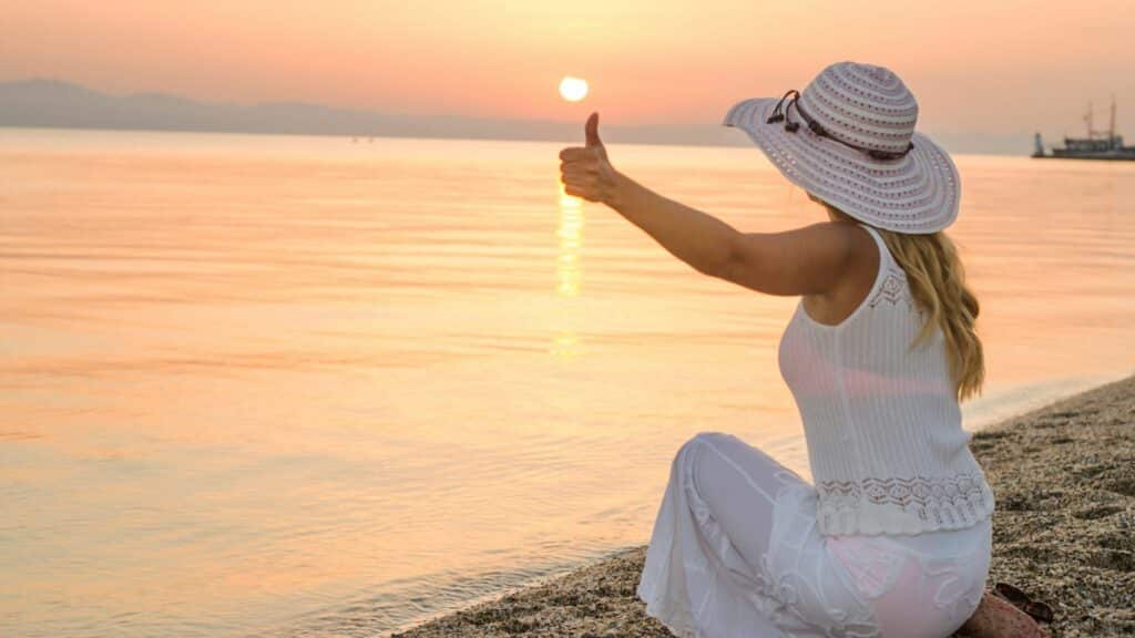 Imagem de uma mulher na praia ao pôr-do-sol fazendo um sinal de positivo com a mão