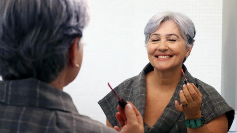 Senhora sorrindo satisfeita para o espelho após fazer sua maquiagem.