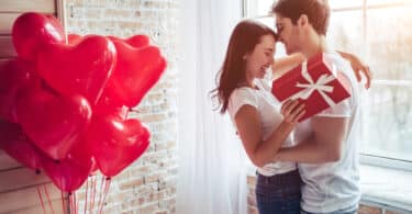 Casal sorridente se abraçando. A mulher segura uma embalagem de presente e, atrás dela, há balões no formato de corações.