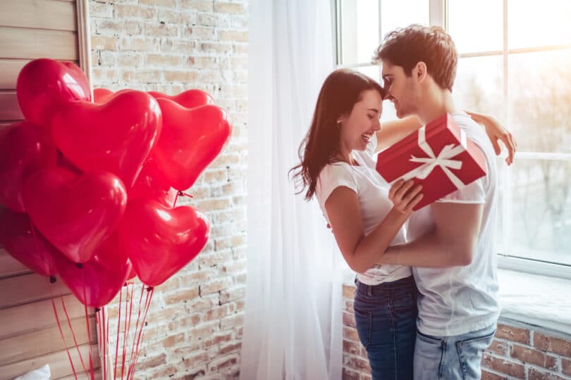 Casal sorridente se abraçando. A mulher segura uma embalagem de presente e, atrás dela, há balões no formato de corações.