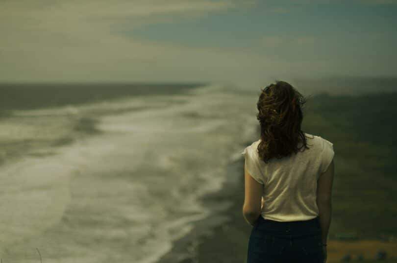 Mulher de costas, na beira do mar, em um dia nublado.