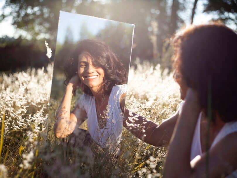 Mulher em frente a um espelho, sorrindo para ele. Ela está em meio à natureza.