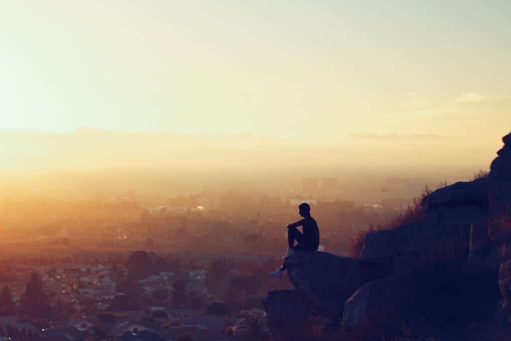 Silhueta de homem sentado na ponta de uma rocha, no alto. Abaixo (e ao fundo) aparece uma cidade. A imagem simboliza sonhar com dinheiro na conta, representando uma vida pacífica.