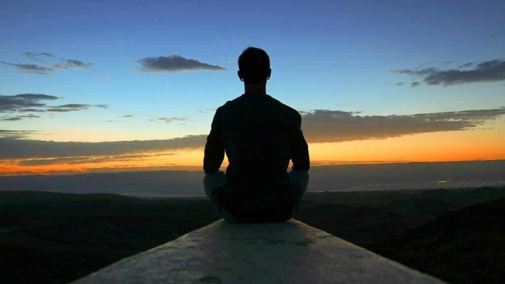 Imagem de um homem sentado de frente para o céu ao final da tarde, como se estivesse meditando