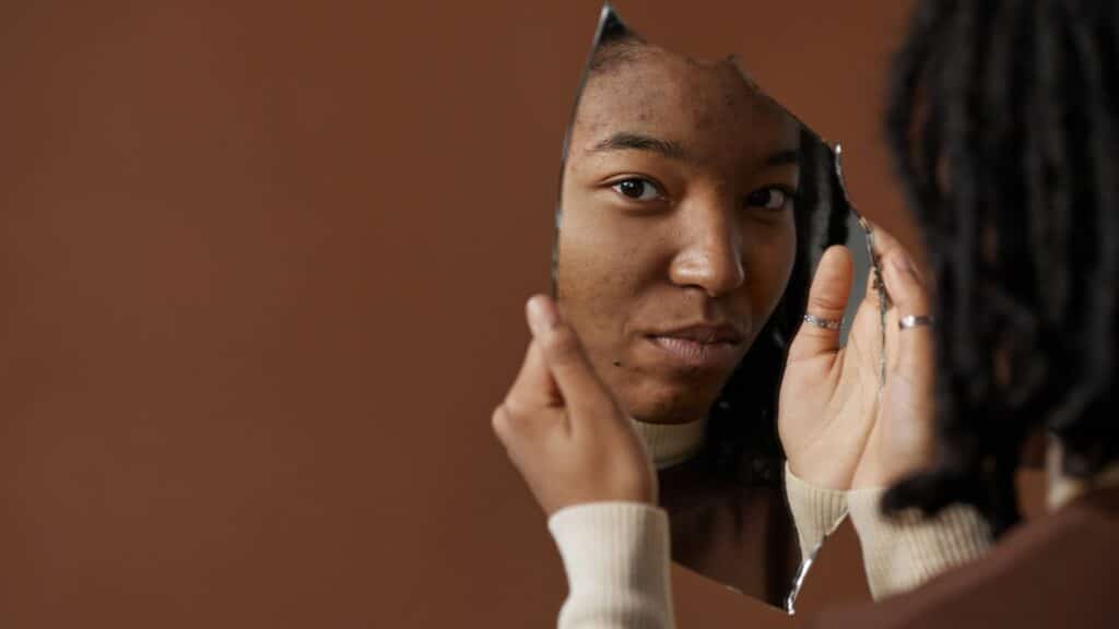 Imagem de uma mulher negra se olhando em um pedaço de espelho em um fundo marrom