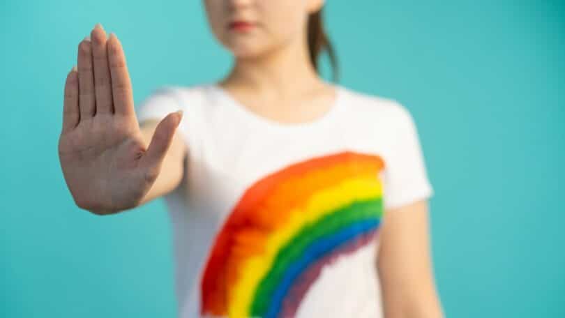 Imagem de uma mulher em um fundo verde com uma roupa branca e um arco-iris desenhado, fazendo sinal de pare com a mão