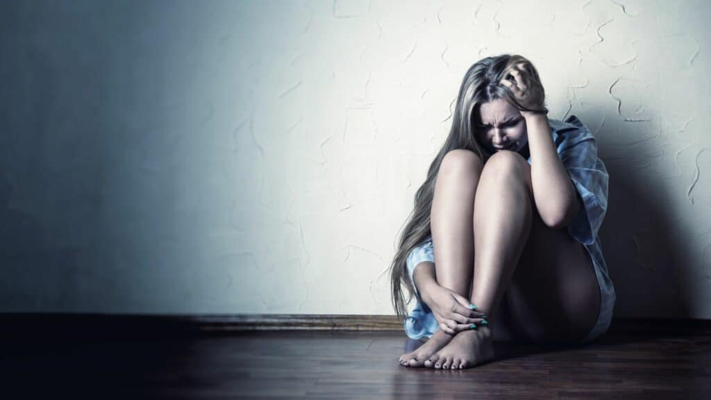 Imagem de uma mulher encostada em uma parede e sentada no chão, chorando e isolada