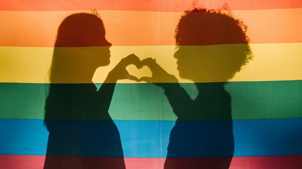 Imagem de duas mulheres atrás de uma bandeira LGBTQIA+ fazendo coração com as mãos