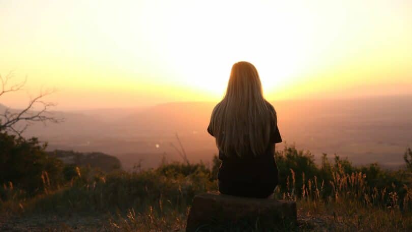 Imagem de uma mulher sentada de costas em uma rocha ao pôr-do-sol