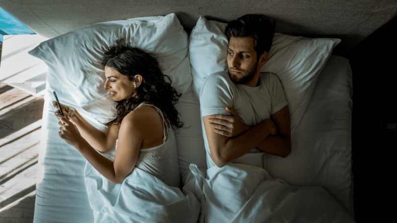 Casal deitado na cama enquanto homem olha para a mulher mexendo no celular