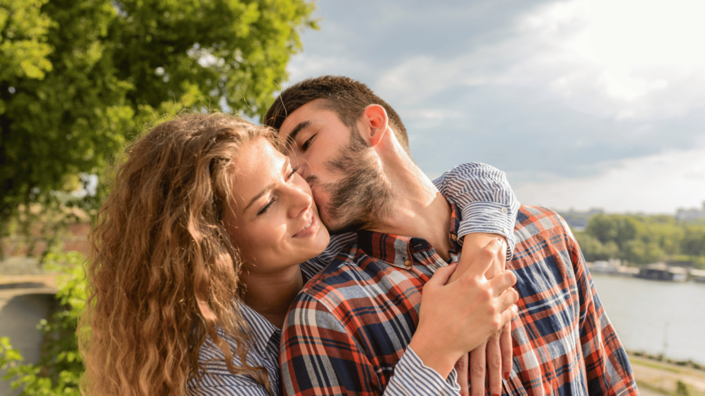 Homem beijando uma mulher loira na bochecha, enquanto ela o abraça.