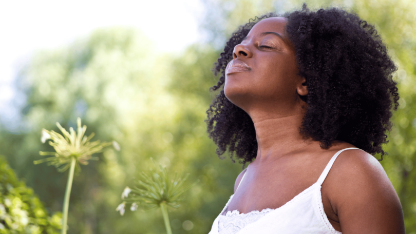 Mulher negra inspirando em um jardim, indicando estar sentindo paz.
